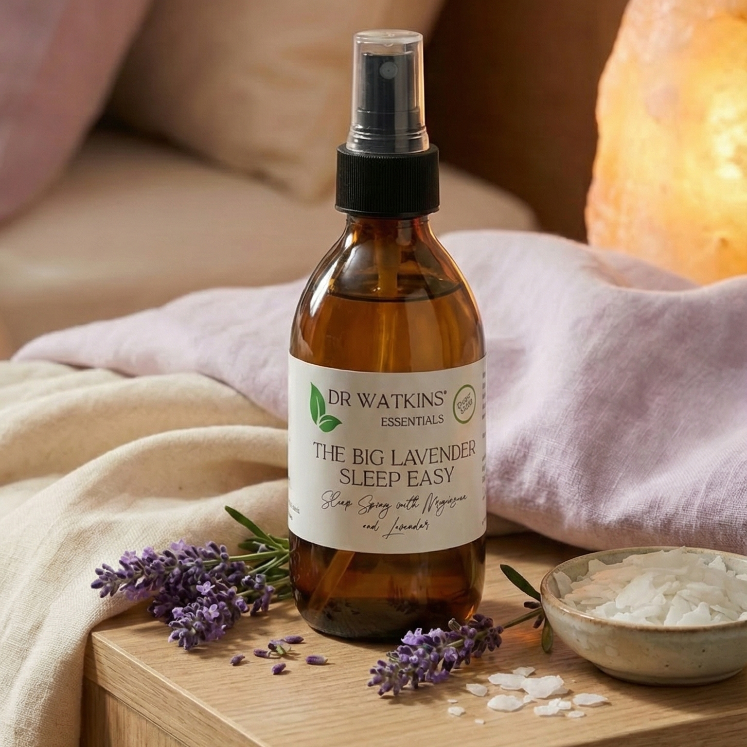 Brown spray bottle labeled 'The Big Lavender Sleep Easy' on a wooden nightstand with lavender flowers and a salt lamp in the background.