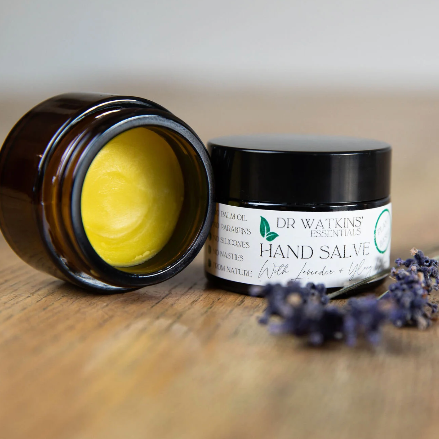 Dr. Watkins Hand Salve jar with a black lid on a wooden surface, accompanied by lavender flowers.